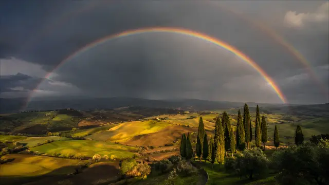 Doble arcoiris sobre paisaje colinoso y cipreses
